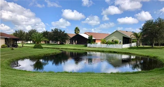 A landscaped residential lot with a small pond in the foreground, green grass, trees, and single-story houses under a blue sky with white clouds.