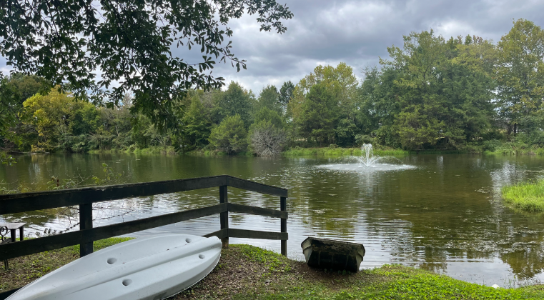 A scenic outdoor view of a calm lake with a fountain in the distance, surrounded by green trees under a cloudy sky. In the foreground, there is a wooden fence, a white boat hull resting on the grass, and a dark container.